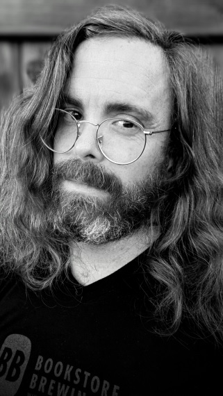 A black and white portrait of Julian Day in front of a wooden fence. He has long, wavy hair, a greying beard, and is wearing a black t-shirt and gold-rimmed glasses.