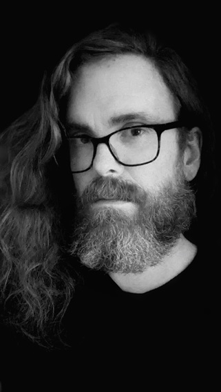 A black and white portrait of Julian Day. He has long, wavy hair, a greying beard, and is wearing glasses and a black t-shirt.